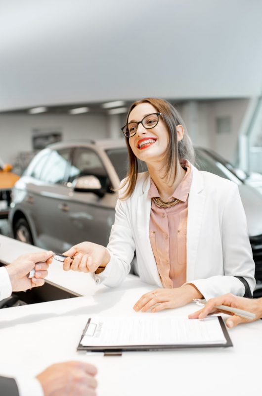 Young business couple signing some documents at the table with salesperson or manager buying or renting car in the showroom